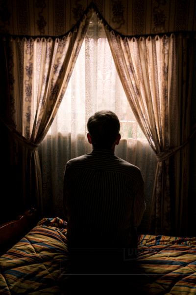 Man sat on bed in darkened room looking glumly out of a veiled window